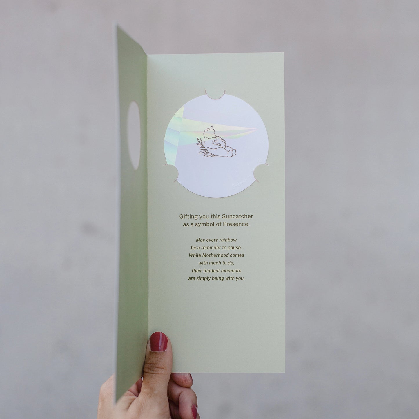 Hand holding a #10 baby card from the Stray Folk SunSpeak Series. The greeting card is open to reveal a light mint green interior. There is a circle suncatcher sticker with a line drawing of a mother bear leaning back with a cub on her belly. The card reads "Gifting you this Suncatcher as a symbol of Presence. May every rainbow be a reminder to pause. While Motherhood comes with much to do, their fondest memories are simply being with you."