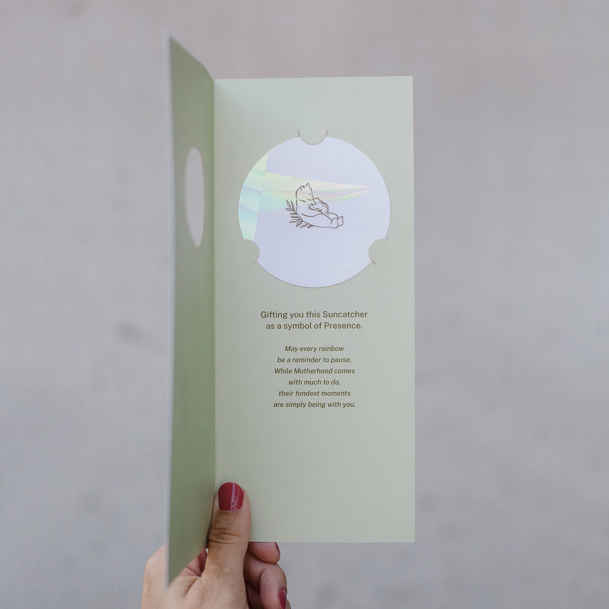 Hand holding a #10 baby card from the Stray Folk SunSpeak Series. The greeting card is open to reveal a light mint green interior. There is a circle suncatcher sticker with a line drawing of a mother bear leaning back with a cub on her belly. The card reads "Gifting you this Suncatcher as a symbol of Presence. May every rainbow be a reminder to pause. While Motherhood comes with much to do, their fondest memories are simply being with you."